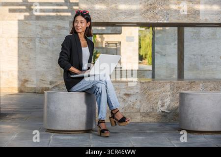 A woman sitting on stone ledge outside with a laptop, dressed casually in a blazer and jeans. Spain Stock Photo