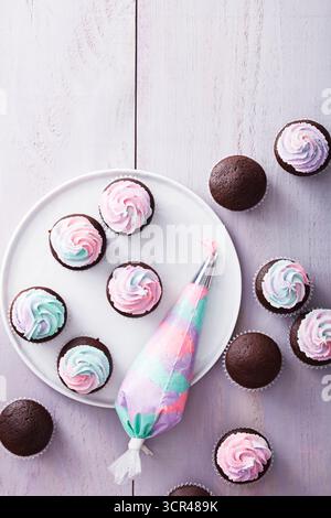 Chocolate cupcakes with pastel swirl frosting arranged on a wooden surface with a piping bag. Stock Photo