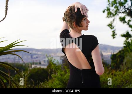 Woman in cutout black dress poses outdoors with city skyline and trees in the background. Stock Photo