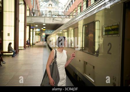 Woman in white dress standing beside a train, looking through the window at a station. Stock Photo