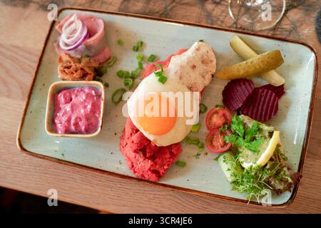 Plated dish with fried egg, pickles, beetroot, and sauce garnished with herbs on a wooden table. North Sea, Germany Stock Photo