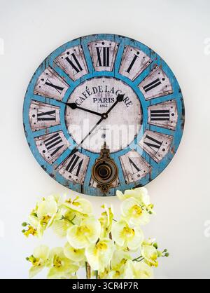 A large blue coloured wall clock with Roman numerals, hanging on a white wall with a bowl of yellow Orchids in front Stock Photo