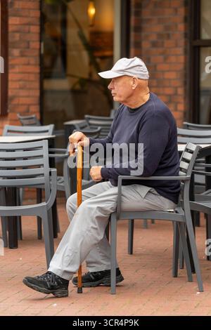 A solitary elderly man wearing a baseball cap and holding a wooden cane, sitting on a green plastic chair outside a cafe in Cleveleys, Lancashire, UK Stock Photo