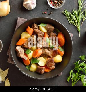 Beef stew with potatoes, carrots and greens in black bowl on dark stone background. Comfort healthy food for dinner. Top view, flat lay Stock Photo