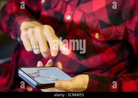 Casual man in red plaid shirt using smartphone indoors displaying a trading app with crypto chart on the screen Stock Photo