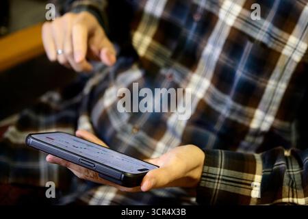 Adult male in plaid shirt seated indoors checking cryptocurrency chart on smartphone screen Stock Photo