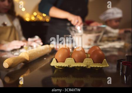 A warm, cozy kitchen scene shows a carton of brown eggs on a reflective countertop, a wooden rolling pin, and blurred bakers in the background, hintin Stock Photo