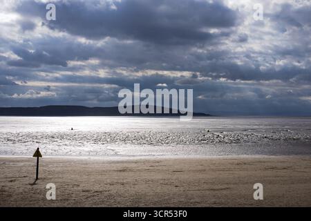 Dramatic clouds over a beach at low tide. A warning sign stands on the sand, cautioning about the mudflats. Stock Photo