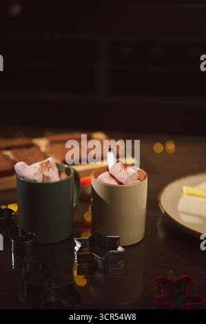 Cozy baking setup featuring mugs with marshmallows, cookie cutters on a dark wooden table, and a plate in the background. Warm, festive mood ideal for Stock Photo