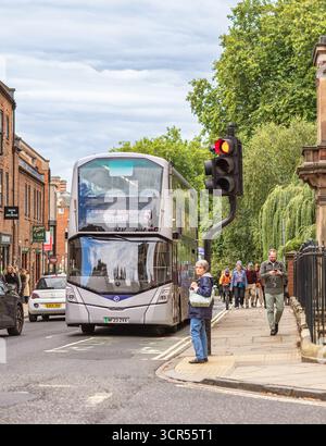A city street corner where a bus waits by a traffic light and pedestrians are on the pavement. Trees  are in the background and a cloudy sky is above. Stock Photo