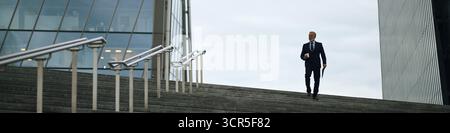 Header of middle aged Caucasian man walking down outdoor steps holding coffee cup and folder, wearing business attire, modern glass building in background, looking ahead with serious expression Stock Photo