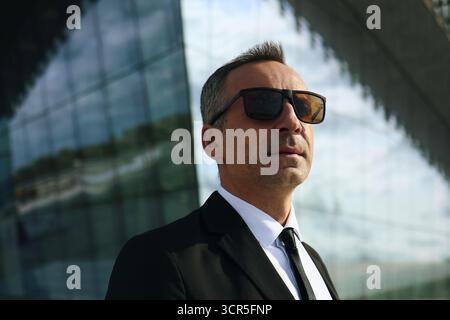 Caucasian middle aged smartly dressed man wearing sunglasses standing outdoors in front of modern glass building looking confidently into distance, business professional posing for corporate concept Stock Photo