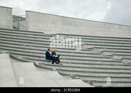 Two Caucasian middle aged business professionals sitting on outdoor concrete steps discussing work while using laptop computer, both focused on screen, urban setting, while taking walk during break Stock Photo
