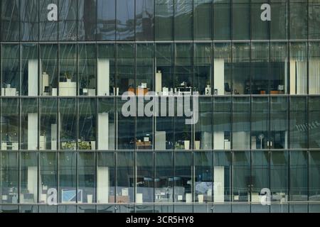 Modern office building exterior showing multiple floors with empty workstations, desks, and office equipment visible through large glass windows, no people present in scene Stock Photo