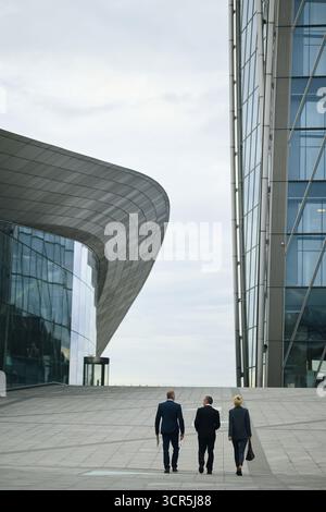 Three middle aged business professionals, two Caucasian men and one woman, walking together outdoors between modern glass office buildings, carrying briefcases, on way to negotiations Stock Photo