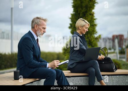 Middle aged Caucasian businessman reading document while sitting outdoors next to female colleague working on laptop, both dressed in business attire, urban background visible Stock Photo