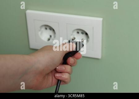 Woman inserting electric plug into power socket on green wall, closeup Stock Photo