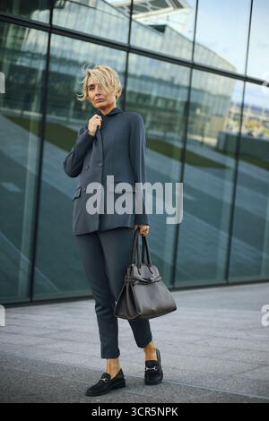 Caucasian middle aged woman walking outdoors holding handbag, short blonde hair, holding jacket collar from wind or chill, glass office building in background, business professional setting Stock Photo