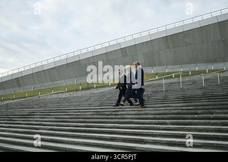 Three middle aged Caucasian corporate colleagues walking down wide outdoor concrete steps in business attire, engaged in conversation, modern architectural structure in background, overcast sky Stock Photo