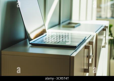 Laptop Office Cabinet Archive Sunlight Interior. Modern open laptop placed on top of a beige filing cabinet in a sunlit office space. The cabinet like Stock Photo