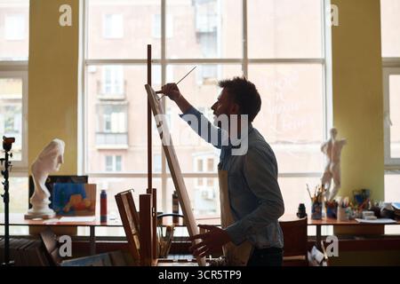 Having art class in studio. Enthusiastic male student in apron finishing his acrylic painting using easel and paintbrush, side view Stock Photo