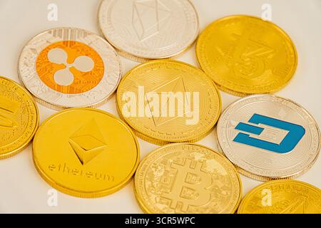 A collection of gold-colored cryptocurrency themed collectible coins scattered on a white surface Coins bear logos of various digital currencies like Stock Photo