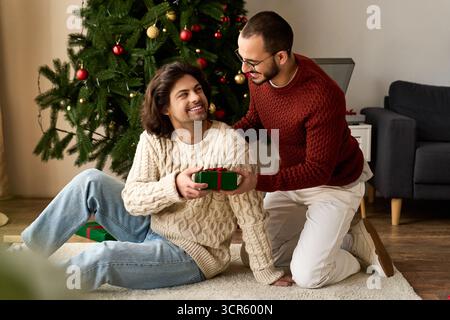 Two men share a heartfelt moment exchanging gifts at home, filled with laughter and warmth. Stock Photo