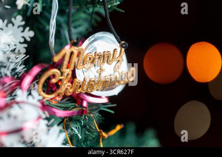 Close-up of golden “Merry Christmas” ornament hanging on a decorated Christmas tree with glowing festive bokeh lights in the background. Stock Photo