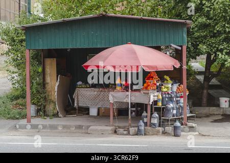 View of a roadside stall, shaded by a red umbrella, displays a colorful array of fruits and drinks in the Armenian countryside, Areni, Vayots Dzor Province, Armenia. Stock Photo