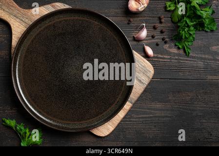 Empty black cast iron platter with herbs and garlic on dark wooden background, top view, copy space. Serving plate mockup. Food background, design tem Stock Photo