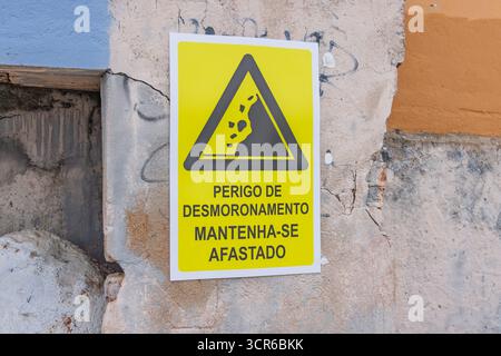 A close-up of a yellow warning sign on a decaying wall in Portugal. The sign shows a falling rock icon and Portuguese text: "PERIGO DE DESMORONAMENTO Stock Photo