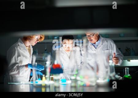 Two students in white coats and protective glasses conducting chemical experiment in university lab under the control of their professor Stock Photo