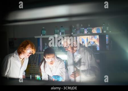 Side view of male and female medical scientists mixing liquids in test tube in order to get new medicine for cancer. Young specialists working under direction of their senior colleague Stock Photo
