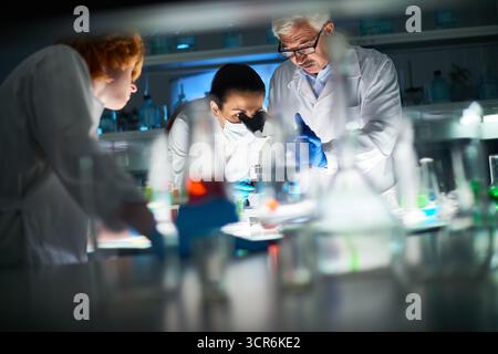 Process of creation new flu shot. Confident scientists in white coats standing at the table with laboratory glassware. Young woman examining chemical substance with the help of microscope. Stock Photo