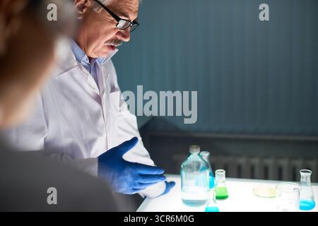 Profile view of senior chemistry teacher in eyeglasses standing at the laboratory table and explaining experiment to his students. Glass bottle with liquid, flasks and Petri dishes located on the desk Stock Photo