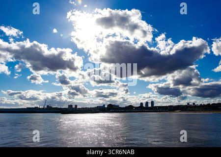 Dramatic sky with scattered clouds over a urban waterfront skyline reflecting sunlight on the water during daytime from Roker Pier Stock Photo