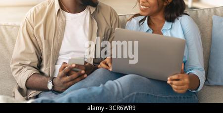 Happy black couple sitting on couch, using cellphone and pc Stock Photo