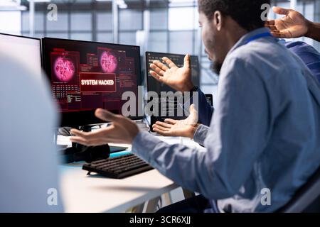 Team of african american engineers in server farm confused by hacker managing to pass firewall and stealing data. Critical error notification on monitor in data center bewildering colleagues Stock Photo