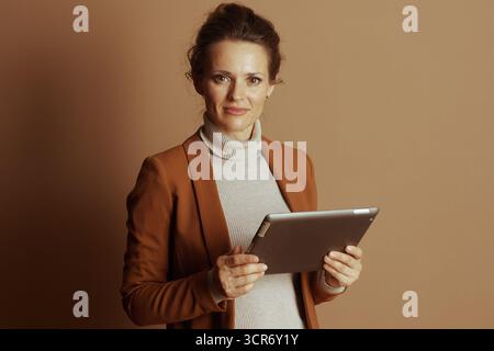 A confident businesswoman with smile holds digital tablet horizontally, looking directly at the camera. Her warm-toned blazer and modern ambiance conv Stock Photo
