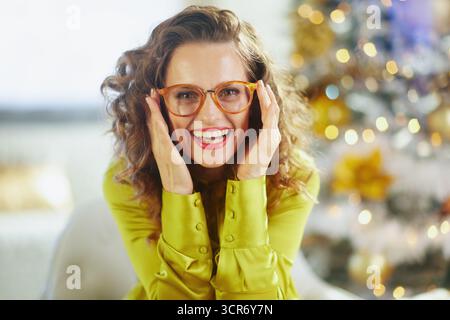 A vibrant woman with curly brown hair and amber glasses smiles, hands framing face. Behind her, blurred Christmas tree glows with warm lights, capturi Stock Photo