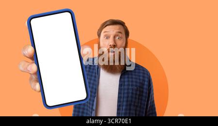 Enthusiastic Man Holding Smartphone Against Orange Background Showcasing a Blank Screen With Surprise Expression Stock Photo