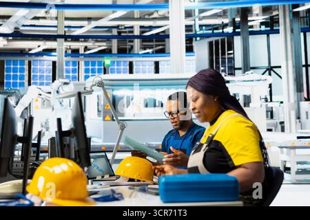 Black engineer in solar panel factory reviewing photovoltaic system performance on laptop. Female employee analyzing production line to optimize efficiency in industrial manufacturing plant. Stock Photo