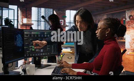 Man in AI startup office CTO position overseeing software engineer implementing machine learning. Artificial intelligence tech venture supervisor helps employee fix code bugs, camera A Stock Photo