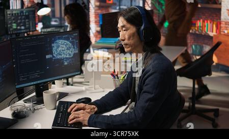 Senior programmer in AI startup office putting on headphones, refining machine learning models to enhance automation. Asian CTO manager listening music while working, camera A Stock Photo