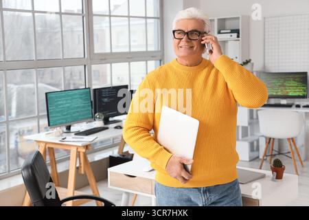 Senior programmer with laptop talking by mobile phone in office Stock Photo
