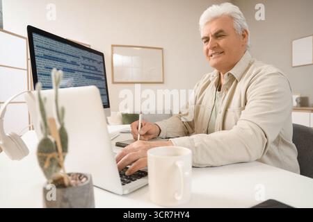 Senior programmer working with laptop and notebook at desk in office Stock Photo