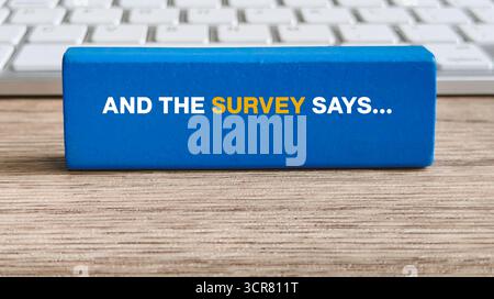 "And The Survey Says..." Text on Blue Wooden Block with Keyboard Background for Research and Feedback Concept Stock Photo