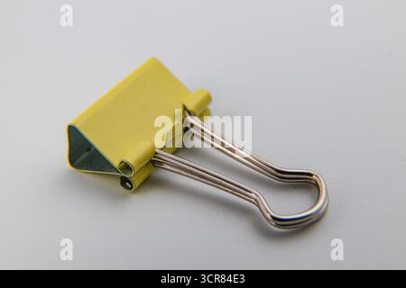 A yellow binder clip is positioned on a smooth white surface, demonstrating its practical design for holding multiple sheets of paper securely togethe Stock Photo
