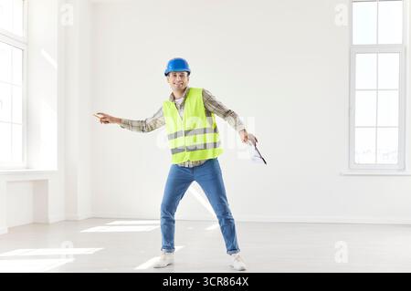 Young happy foreman working in empty new apartment wearing uniform and helmet. Stock Photo