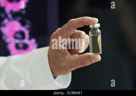 Hand of anonymous male researcher holding small glass vial filled with yellow liquid between fingers, laboratory background with scientific imagery visible, focus on hand and vial Stock Photo